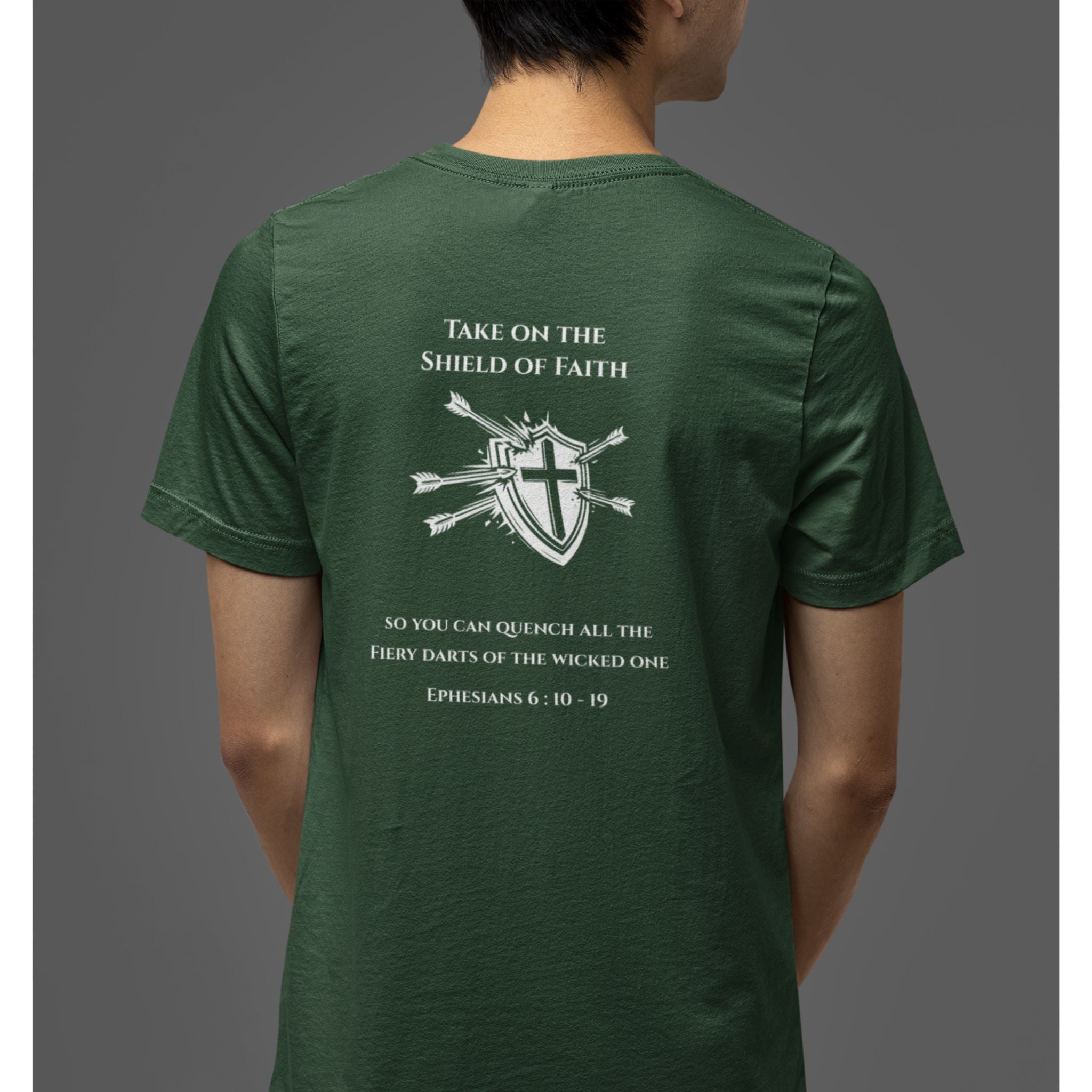 A man wearing a dark green t-shirt, viewed from the back. The shirt features a white graphic print with a shield, cross, arrows, and Bible verse text from Ephesians 6:10-19.