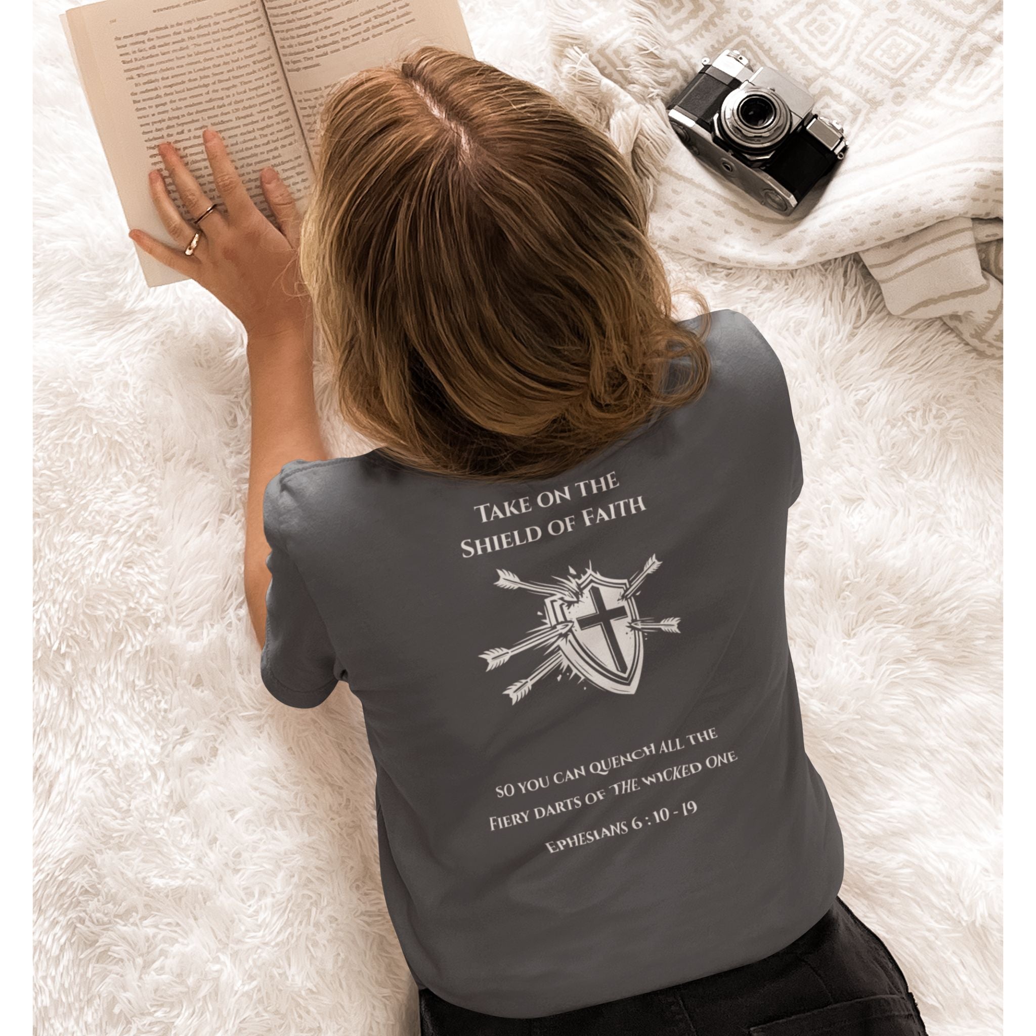 A woman lying on a white shag rug wearing a dark gray t-shirt with a white printed graphic and Bible verse on the back. She is holding an open book, with a vintage camera and a patterned blanket nearby.