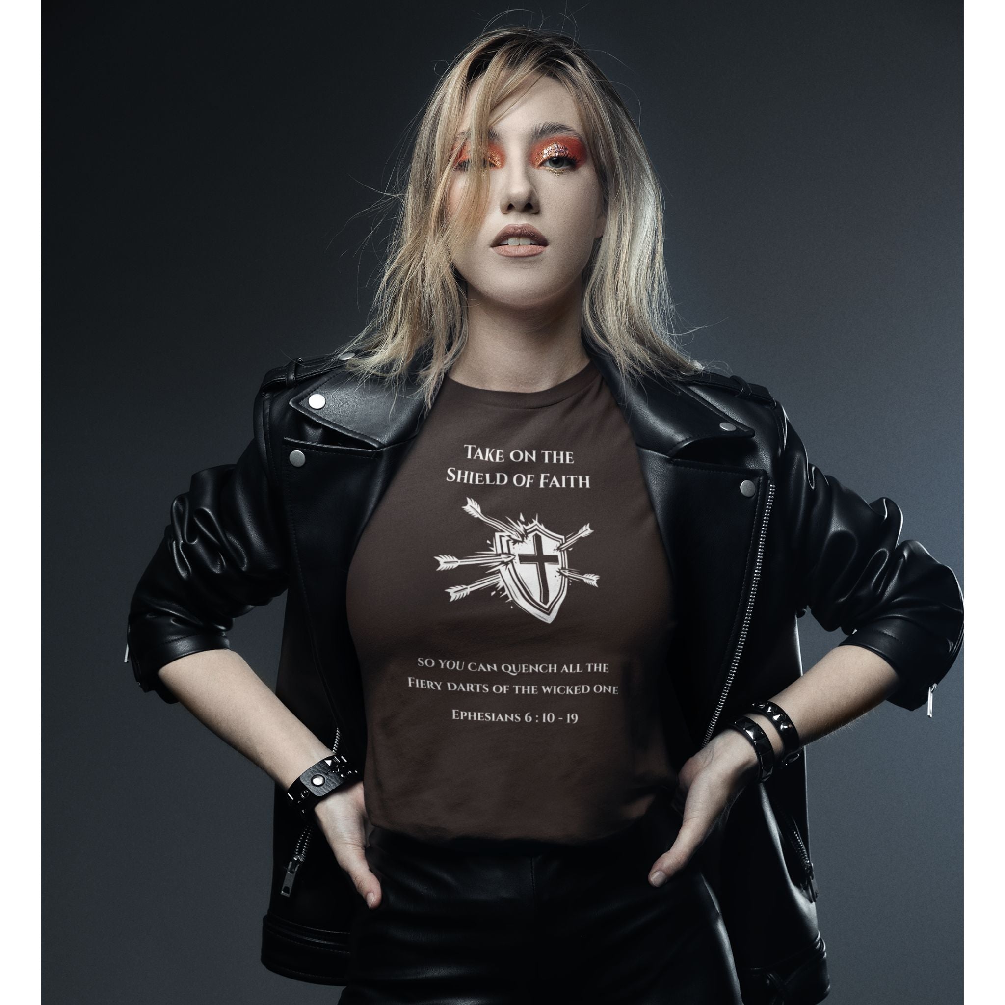 A woman posing in a black leather jacket over a brown graphic t-shirt with a cross and bible verse print, set against a dark gray studio background.