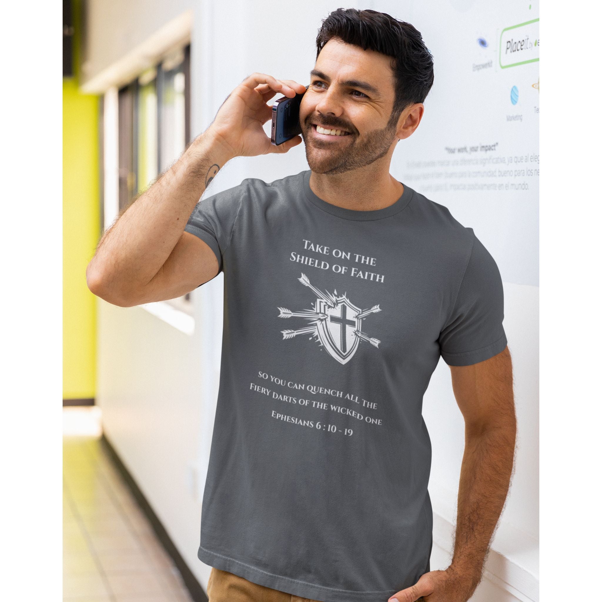 A man wearing a dark gray graphic t-shirt with a white cross and bible verse print, standing in a brightly lit hallway and talking on a mobile phone.