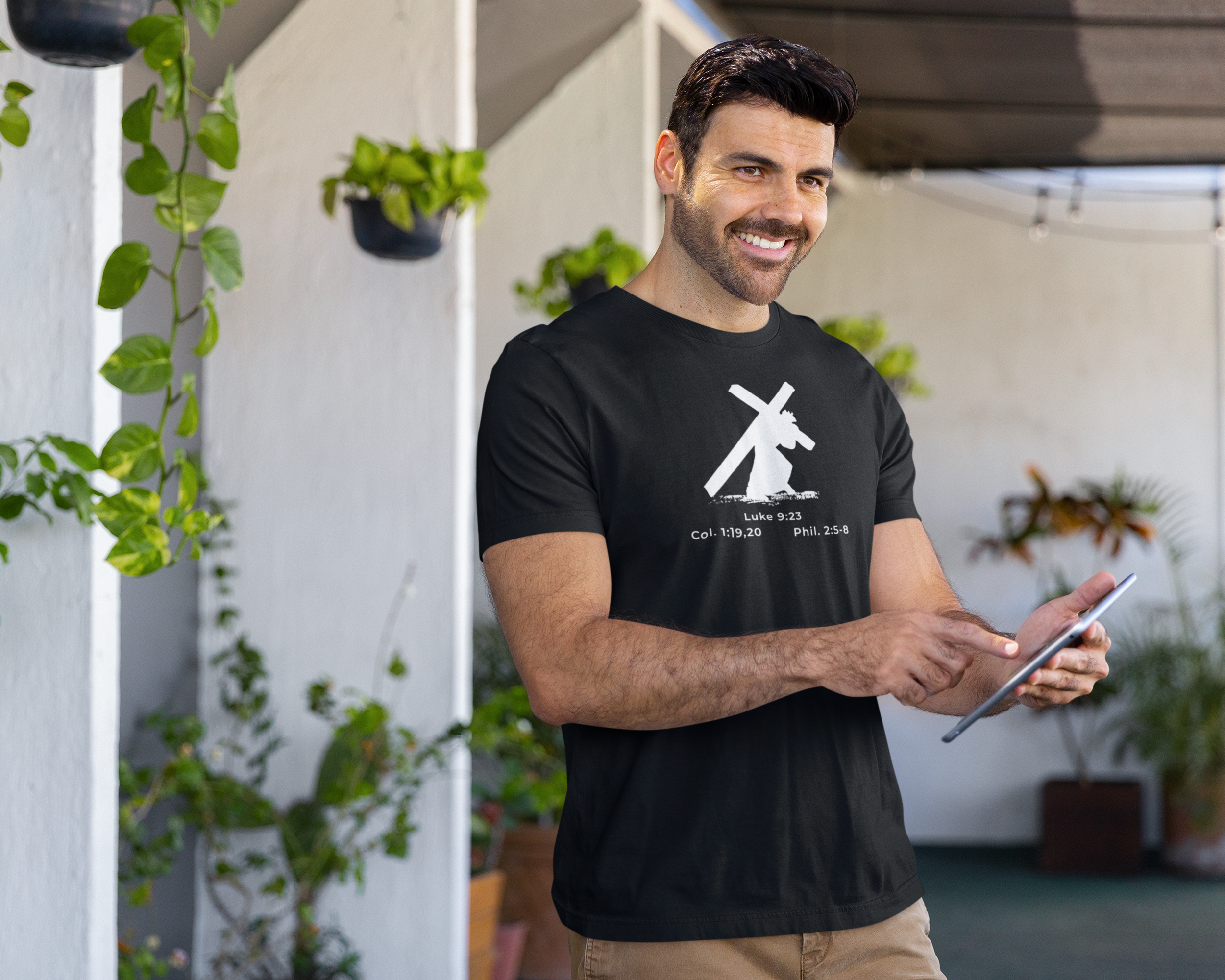 Man wearing a black t-shirt with a white graphic design, holding a tablet, standing in an outdoor setting with plants.