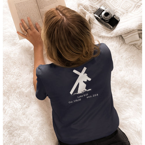 Person wearing a navy blue t-shirt with a cross design and text, lying on a white surface with an open book and camera.