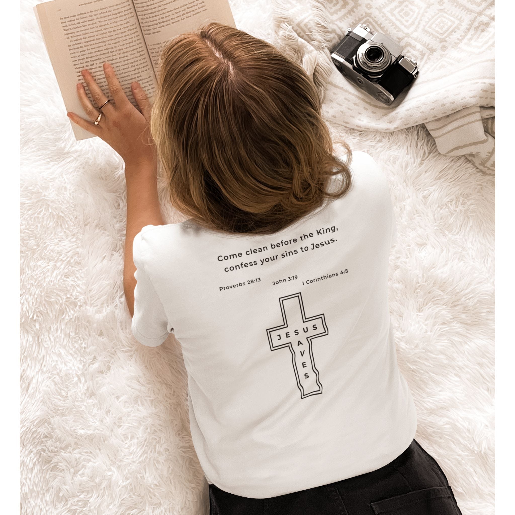 Person wearing a white t-shirt with a cross design and text, lying on a fluffy white surface with an open book and camera.