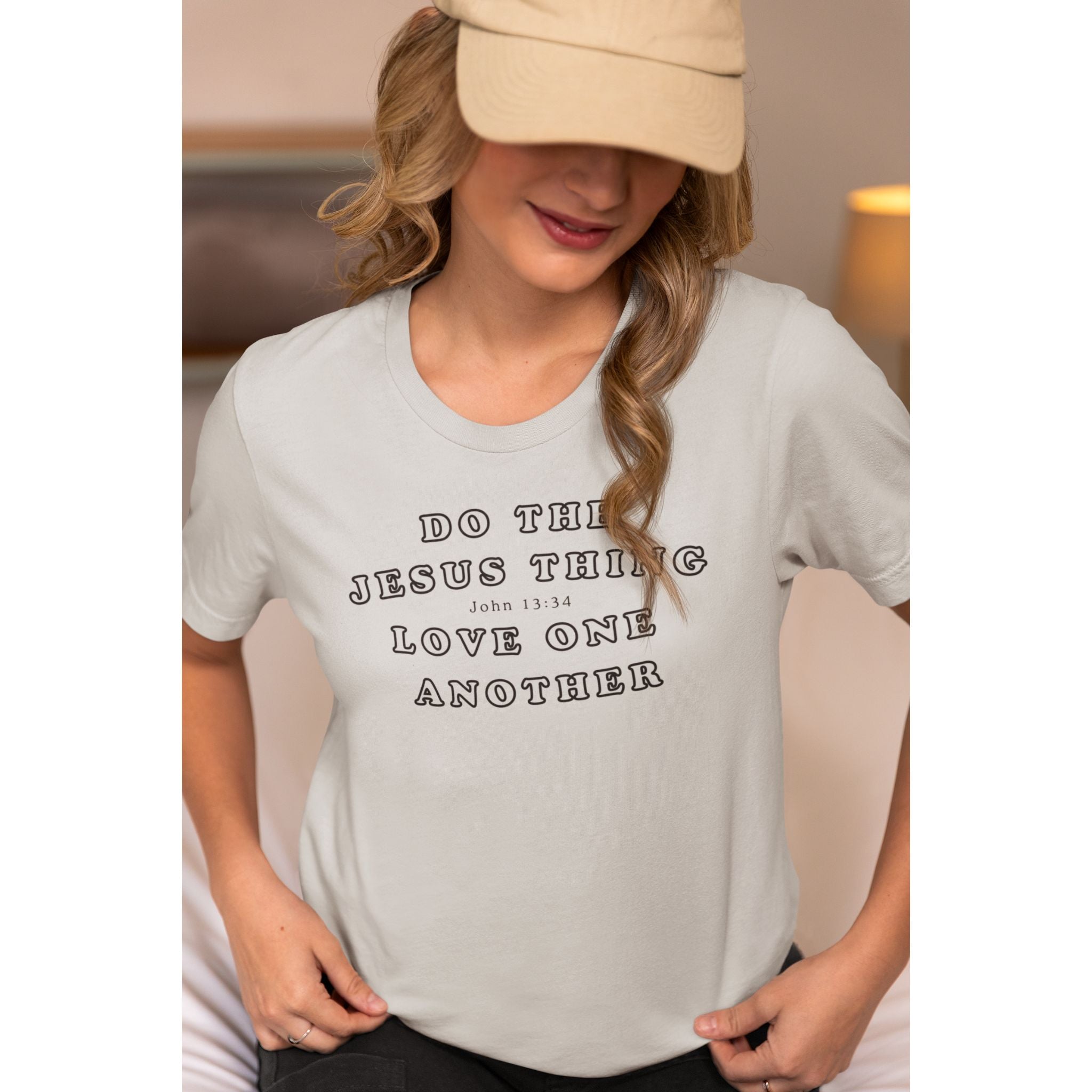 A woman wearing a light beige baseball cap and a light gray graphic t-shirt with the text 'DO THE JESUS THING LOVE ONE ANOTHER John 13:34' printed in black.
