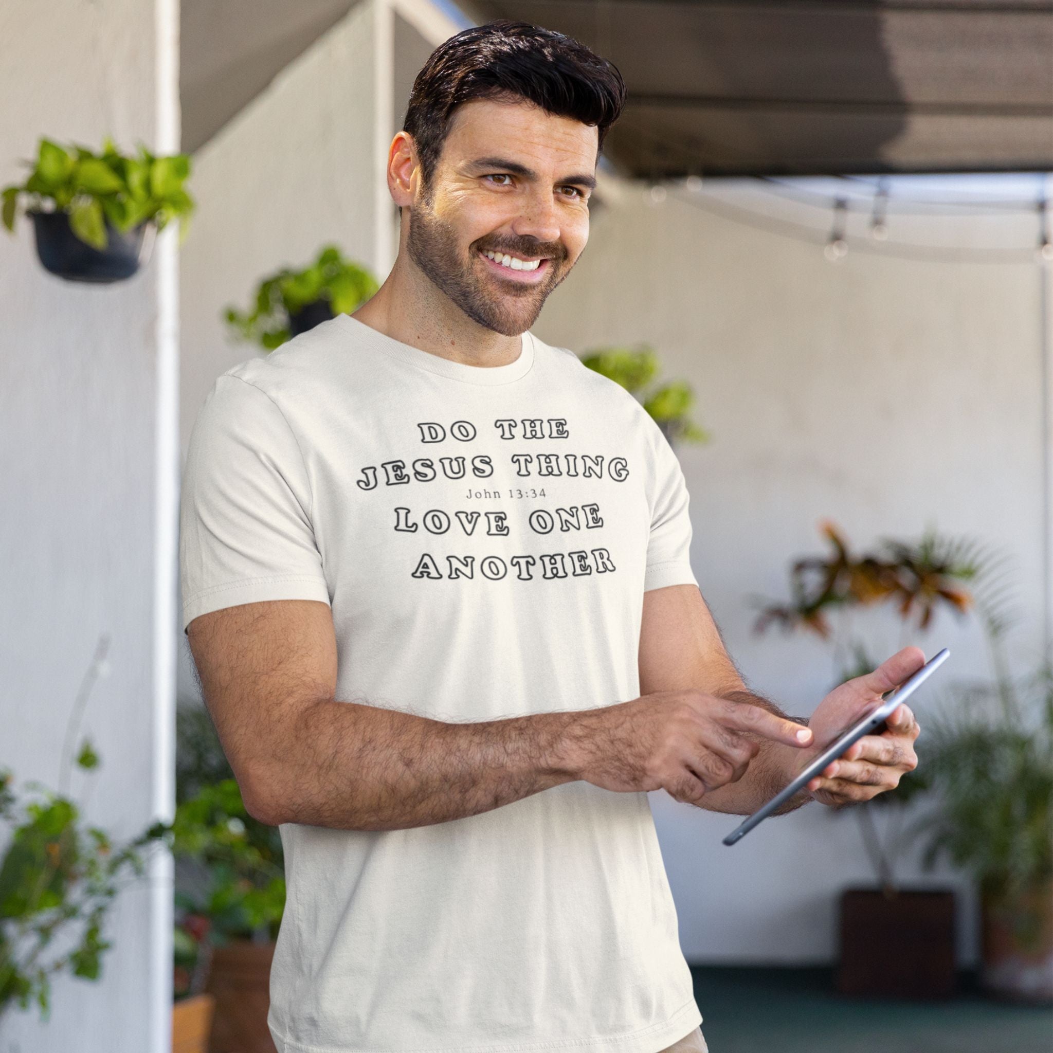 A smiling man wearing a cream-colored t-shirt with the text 'DO THE JESUS THING LOVE ONE ANOTHER' printed in black. He is holding a tablet in a modern, sunlit outdoor patio setting with potted plants in the background.