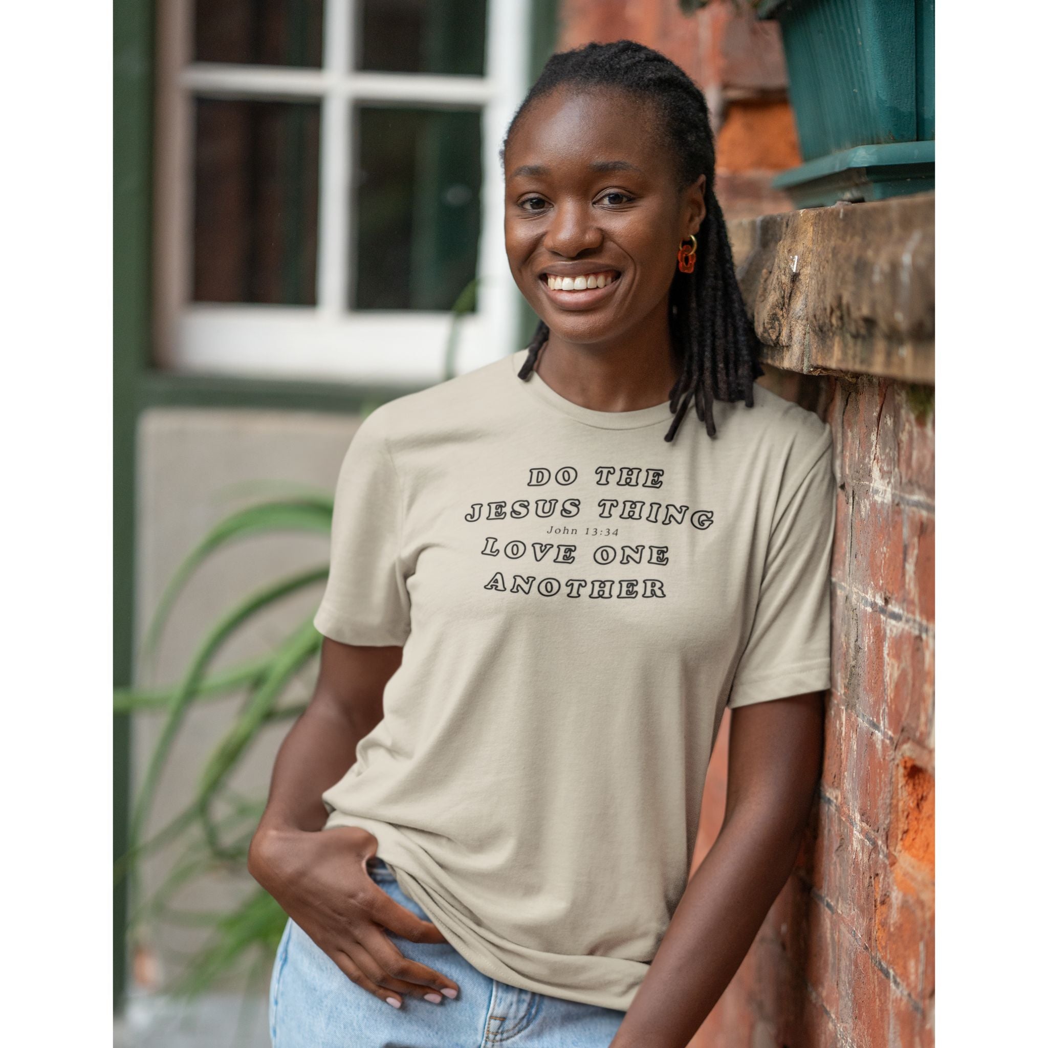 A woman smiling and leaning against a brick wall, wearing a light beige graphic t-shirt with the text 'DO THE JESUS THING LOVE ONE ANOTHER' printed in black.