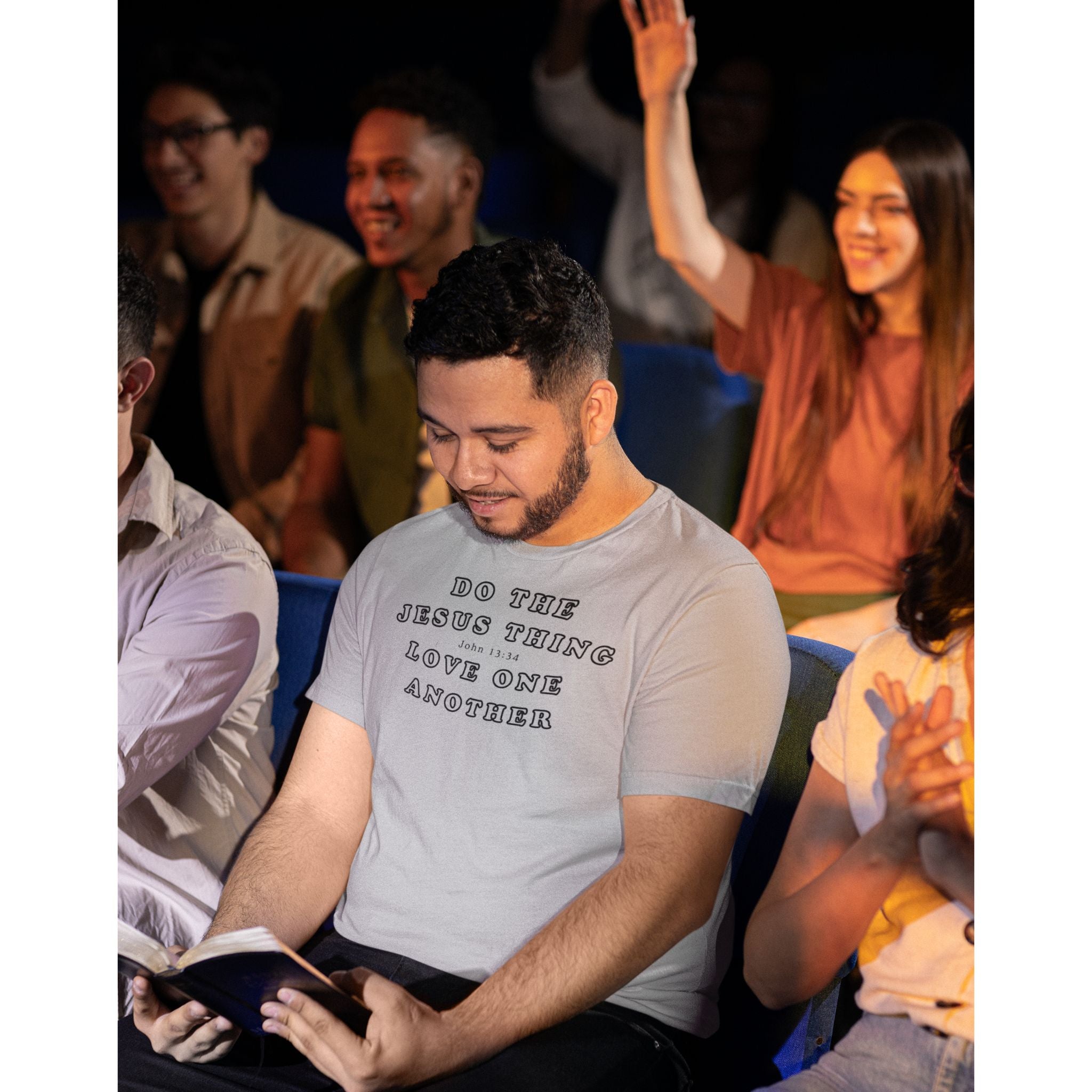 A man in a gray graphic t-shirt sitting in a church setting, reading a Bible. The t-shirt features the text 'DO THE JESUS THING LOVE ONE ANOTHER'.